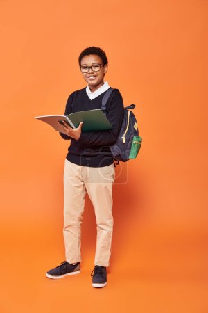 cheerful african american schoolboy in uniform holding backpack and textbook on orange backdrop magic mug #692619090