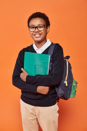 Foto de Relajado afroamericano colegial en uniforme celebración mochila y libro de texto en naranja telón de fondo - Imagen libre de derechos