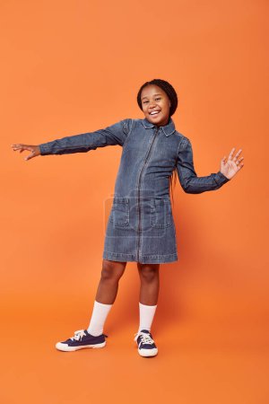 full length of excited african american girl with braids posing in denim dress on orange background