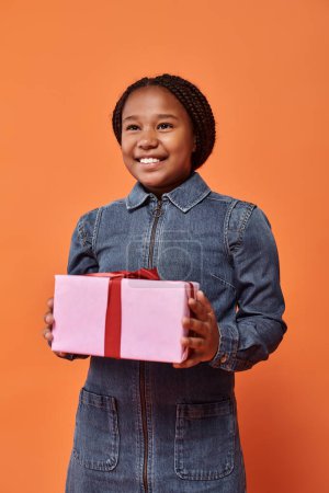 joyful african american girl in denim dress holding wrapped present and looking at camera on orange