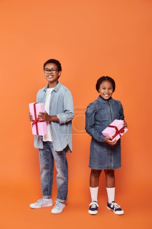 niños afroamericanos felices sosteniendo regalos envueltos y de pie sobre fondo naranja