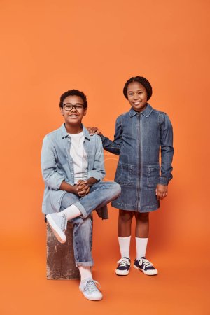 fröhliche afrikanisch-amerikanische Kinder in lässiger Jeans-Kleidung posieren gemeinsam auf orangefarbenem Hintergrund
