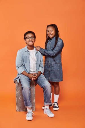 fröhliche afrikanisch-amerikanische Kinder in lässiger Jeans-Kleidung posieren gemeinsam auf orangefarbenem Hintergrund