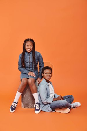 fröhliche afrikanisch-amerikanische Kinder in lässiger Jeanskleidung sitzen zusammen auf orangefarbenem Hintergrund
