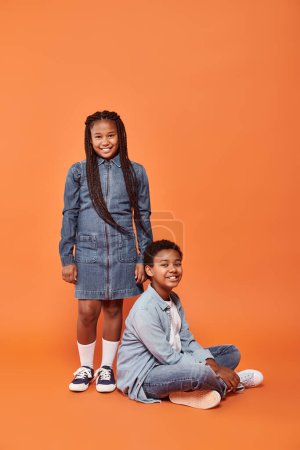 fröhliches afrikanisch-amerikanisches Mädchen in lässiger Jeanskleidung, das neben einem Jungen auf orangefarbenem Hintergrund steht