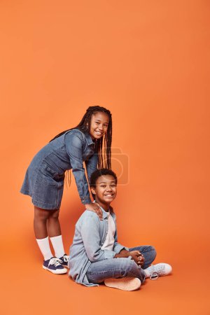 cheerful african american girl in casual denim attire standing and leaning on boy on orange