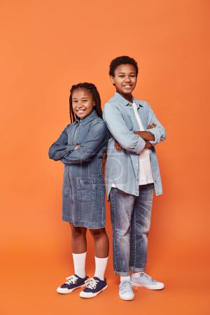 happy african american children in casual denim attire standing with folded arms on orange backdrop