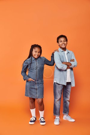 optimistische afrikanisch-amerikanische Kinder in lässiger Jeanskleidung posieren gemeinsam auf orangefarbenem Hintergrund