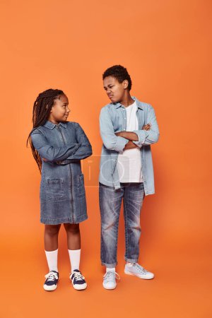 Afrikanisch-amerikanische Kinder in lässiger Jeans-Kleidung posieren mit verschränkten Armen auf orangefarbenem Hintergrund