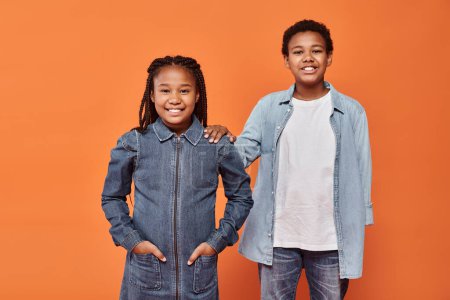 happy african american girl in casual denim attire posing together with boy on orange background