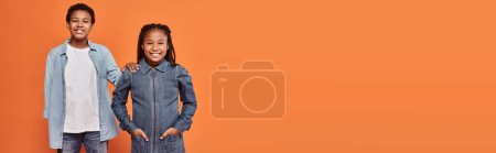 cheerful african american girl in casual denim attire posing together with boy on orange, banner