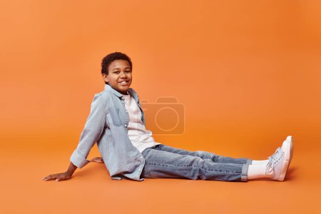 Photo for Joyous preteen arican american boy in everyday attire sitting on floor and smiling at camera - Royalty Free Image
