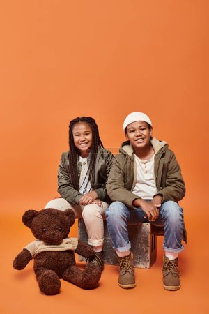 cheerful african american children in warm outfits posing with teddy bear on orange backdrop