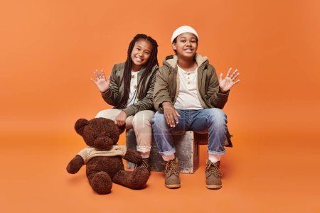 jolly preteen african american children in winter outfit posing with teddy bear and waving at camera