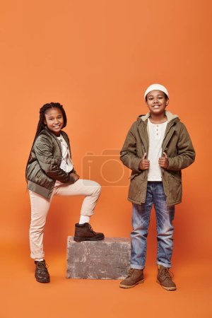 alegre afroamericano niños en invierno trajes posando en ornage telón de fondo y sonriendo felizmente