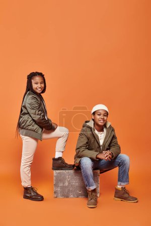 preteen cheerful african american friends in winter outfits posing together on orange backdrop