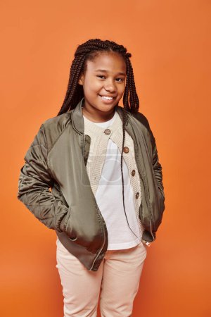 merry preadolescent african american girl with braids posing with hands in pockets smiling at camera