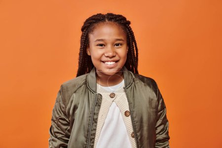 fröhliches afrikanisch-amerikanisches Mädchen in Winterkleidung lächelt freudig in die Kamera auf orangefarbenem Hintergrund
