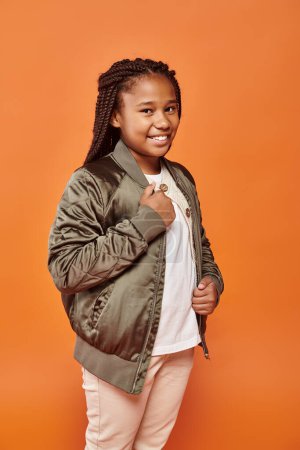 cheerful preteen african american girl in winter outfit smiling happily at camera on orange backdrop