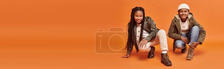 jolly preteen african american children in winter jackets squatting on orange backdrop, banner