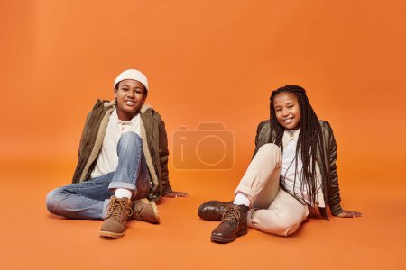 Foto de Alegre preadolescente afroamericano amigos en caliente trajes sentado en el suelo y sonriendo felizmente - Imagen libre de derechos