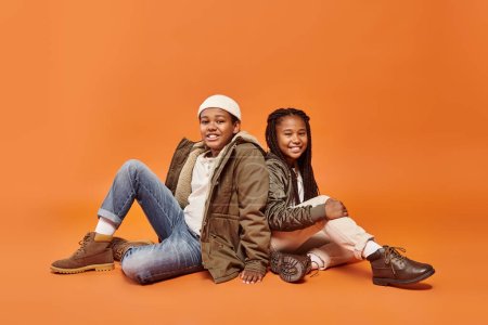 Téléchargez les photos : Joyeux préadolescent afro-américain enfants en tenues d'hiver assis sur le sol souriant à la caméra - en image libre de droit