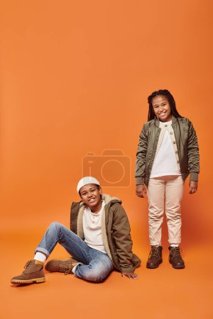 joyous preteen african american children in winter attire smiling at camera on orange background