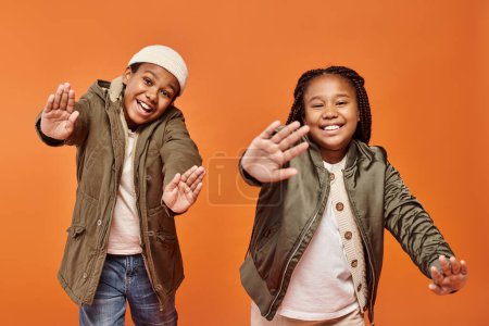 Téléchargez les photos : Joyeuse afro-américaine fille et garçon en vestes d'hiver chaudes posant en mouvement sur fond orange - en image libre de droit