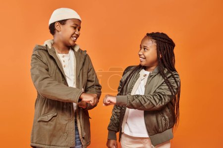 fröhliche afrikanisch-amerikanische Kinder in warmen Winterreifen, die ihre Fäuste auf orangefarbenem Hintergrund schlagen
