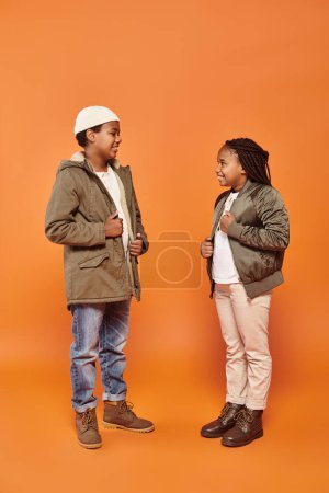 cheerful preteen african american friends in winter attires smiling at each other on orange backdrop