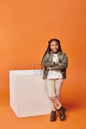 jolly african american girl in warm outfit posing next to white cube with arms crossed on chest