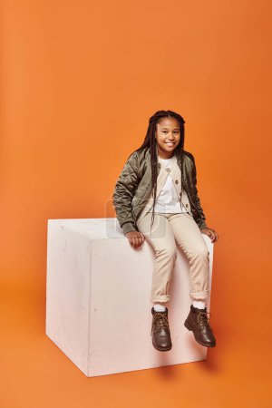 happy african american girl in winter attire smiling at camera on white cube on orange backdrop