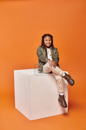 cheerful preteen african american girl in winter attire sitting on cube and smiling at camera