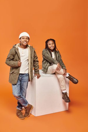happy preteen african american friends in winter outfits smiling ta camera next to white cube
