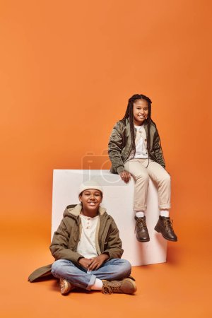 cheerful african american boy and girl smiling at camera next to white cube on orange backdrop