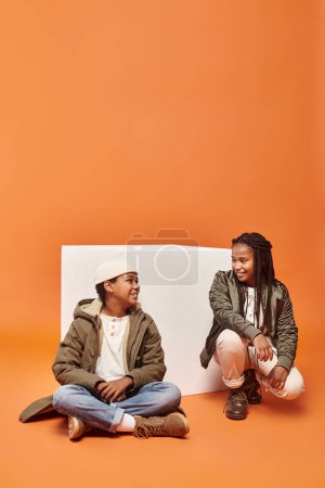 Foto de Alegre afroamericano chica y chico en invierno trajes sentado cerca blanco cubo sonriendo el uno al otro - Imagen libre de derechos