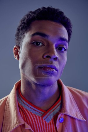 retrato de hombre afroamericano guapo en camisa de melocotón mirando a la cámara en gris con luz azul