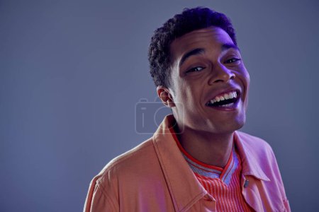 portrait of joyous african american guy in peach shirt laughing on grey background with blue light