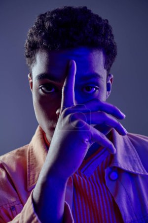 african american man in peach shirt with finger near face looking at camera on grey with blue light