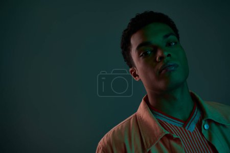 guapo afroamericano hombre en camisa mirando cámara en oscuro estudio con luz verde