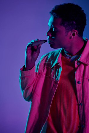 Photo for African american guy vaping with electronic cigarette in hand, grey backdrop with lighting - Royalty Free Image