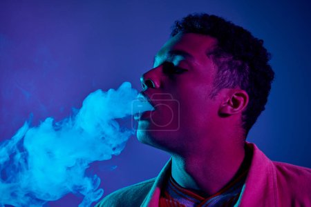 joven afroamericano hombre exhalando humo sobre un fondo azul con iluminación púrpura