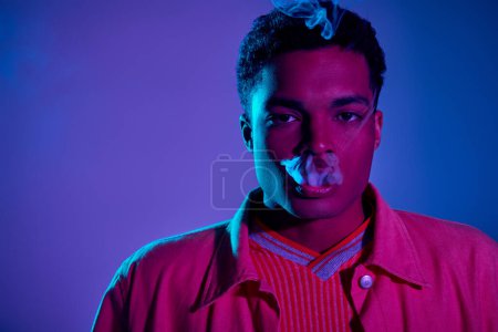 Téléchargez les photos : Jeune homme afro-américain expirant la fumée sur un fond bleu avec un éclairage violet, gen z - en image libre de droit