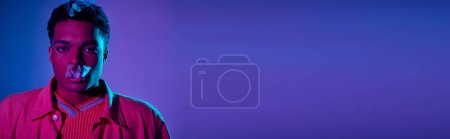 joven afroamericano hombre exhalando humo contra un fondo azul con iluminación púrpura, pancarta