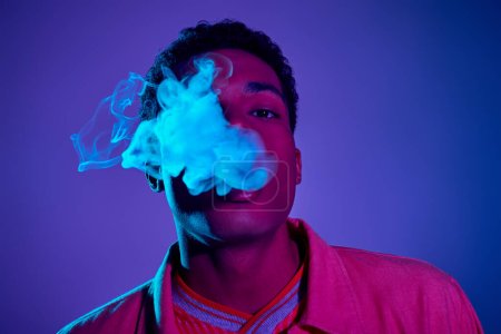 junger afrikanisch-amerikanischer Mann atmet Rauch vor blauem Hintergrund mit violettem Licht ein,