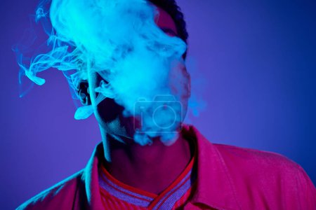 bel homme afro-américain expirant fumée sur fond bleu avec éclairage violet, gen z