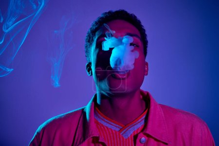 Foto de Tipo afroamericano con estilo exhalando humo contra fondo azul con iluminación púrpura, gen z - Imagen libre de derechos
