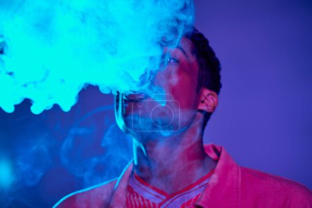 Porträt eines Afroamerikaners, der Rauch vor blauem Hintergrund mit violettem Licht ausatmet, Gen z