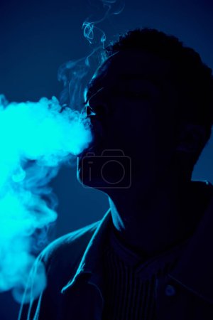 retrato del hombre afroamericano exhalando humo contra fondo oscuro con luz azul, vapeo