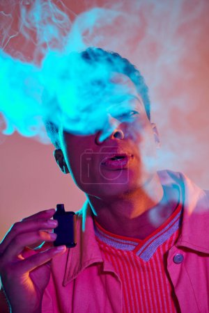 Porträt eines Afroamerikaners, der Rauch ausatmet, während er E-Zigarette vor lebendigem Hintergrund hält